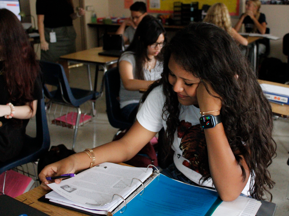 A student with dark hair writing in a notebook, seated in a classroom with others working at desks.