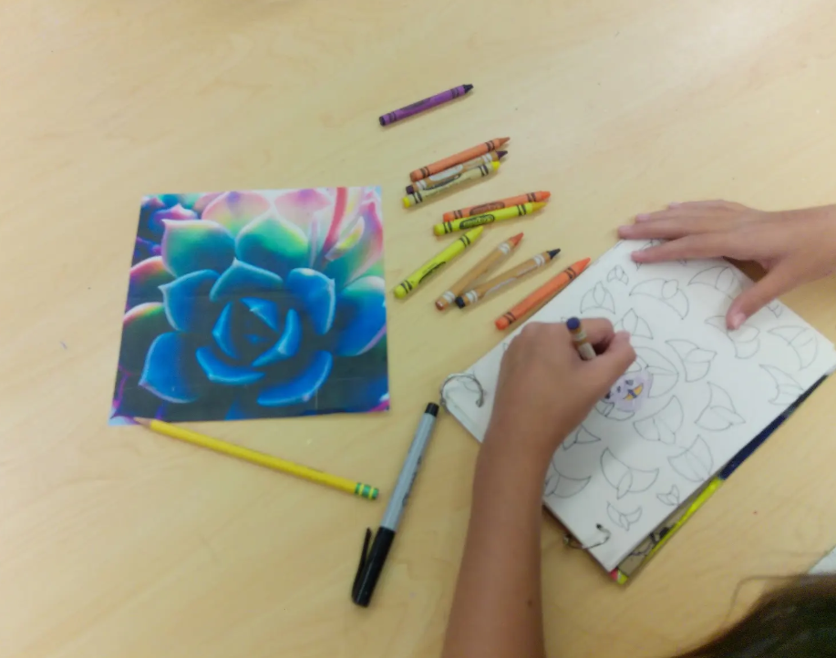 Child coloring a flower on a notebook with colored pencils on a light-colored table.