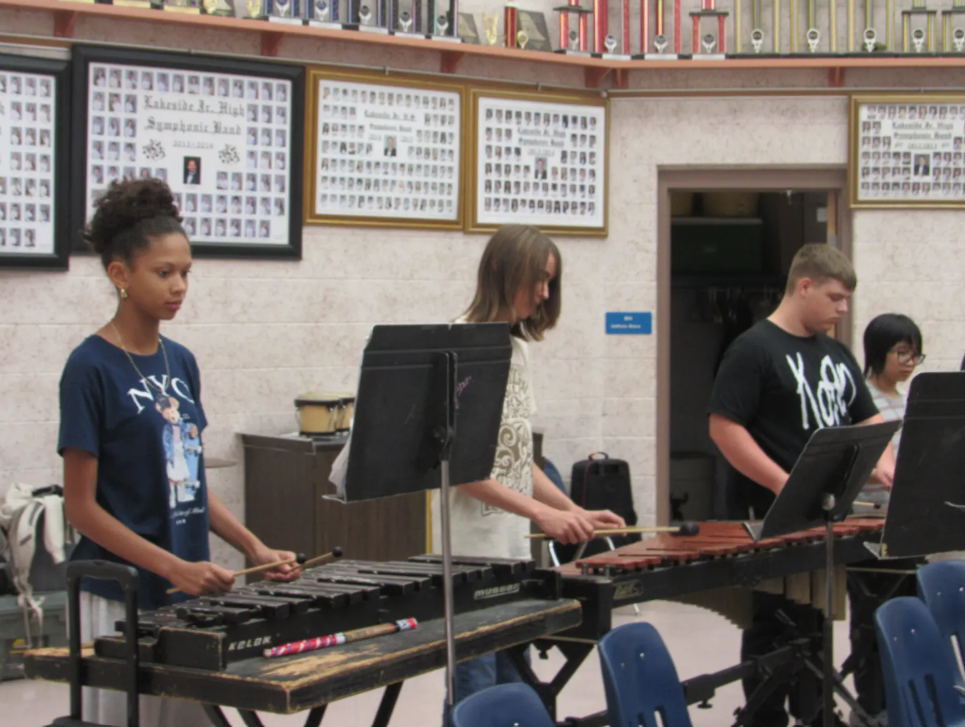 Four students in a classroom with musical instruments; two use xylophones, one uses a marimba, and one plays a piano.
