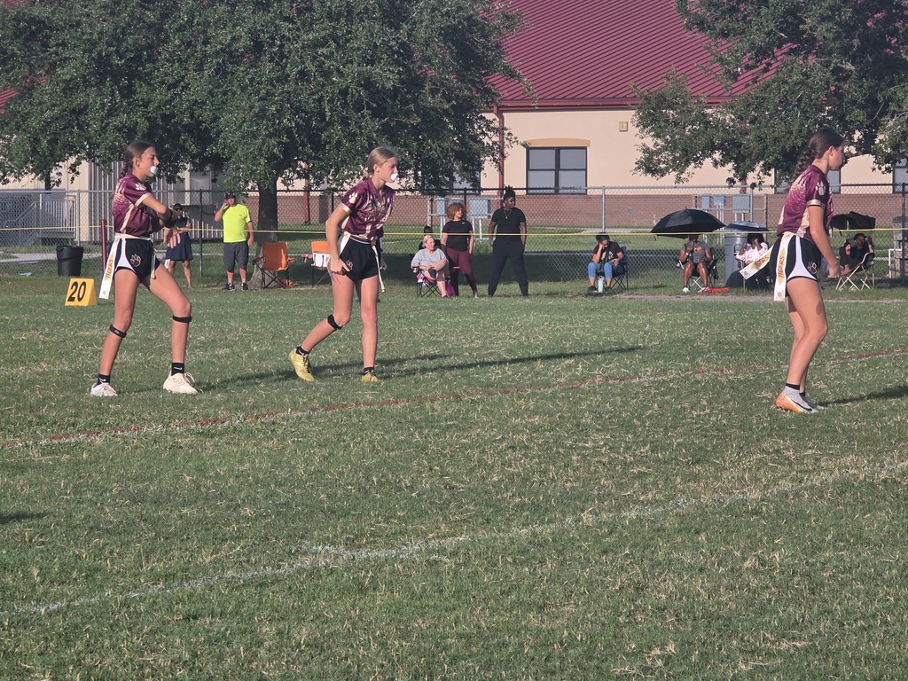 Three people in maroon uniforms play soccer on a field with green grass, trees, and a building.