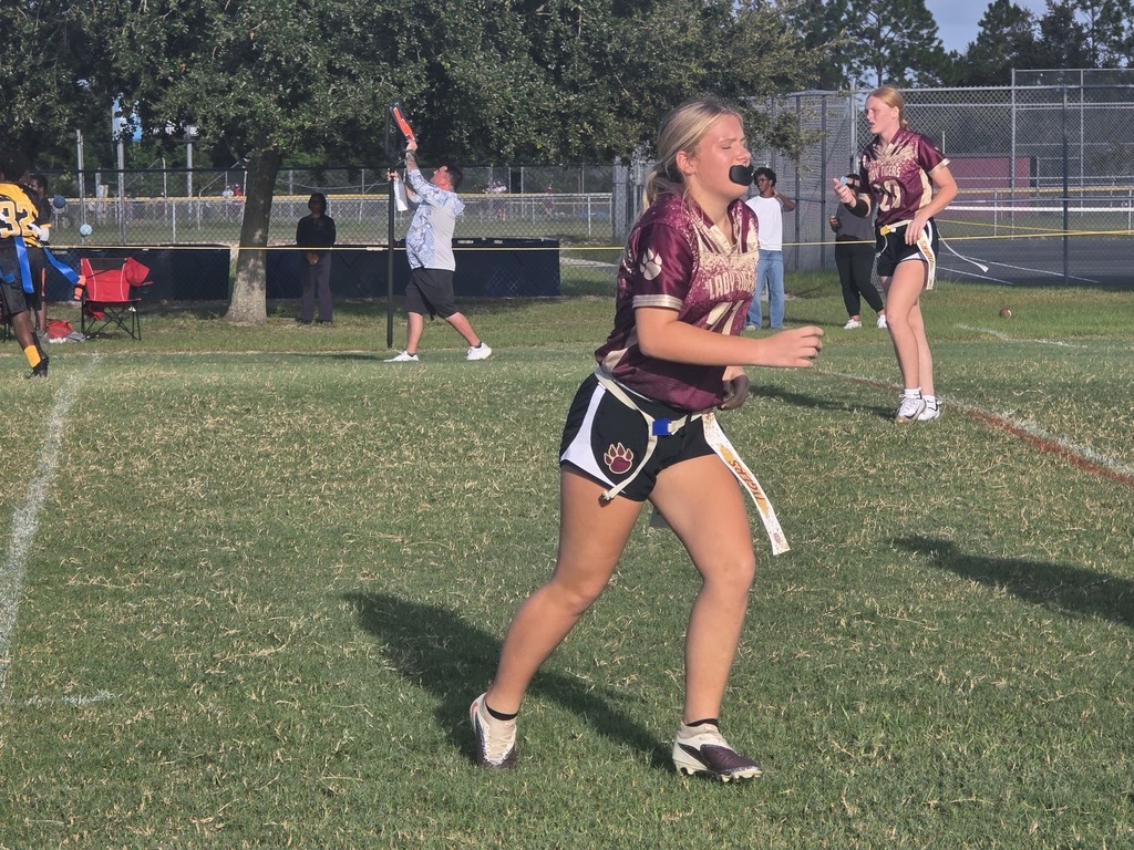 A woman running on a field with other people, holding a football. Trees and a fence in the background.