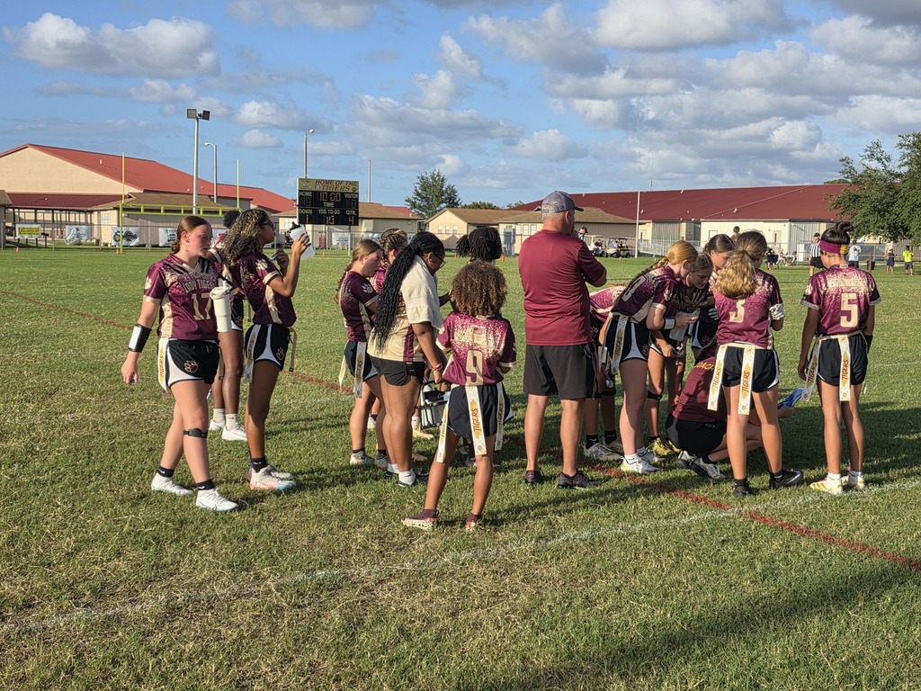 A team gathers on a grassy field, with a coach in a maroon shirt, during a break.
