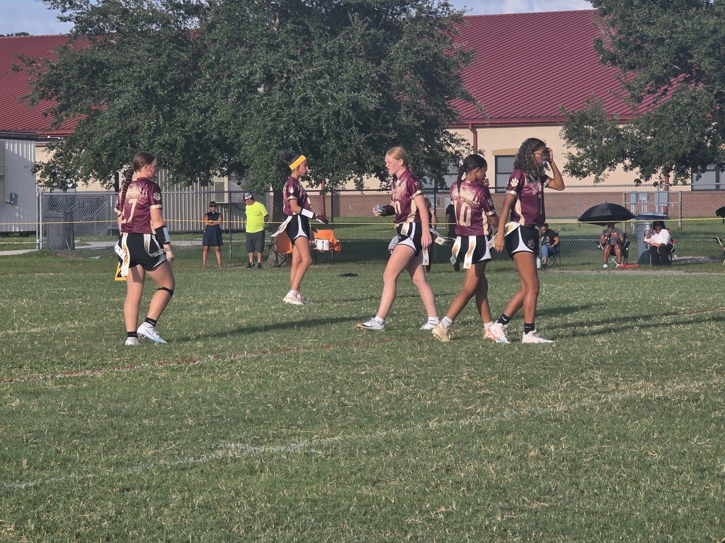 A group of cheerleaders wearing maroon uniforms run on a grass field with buildings in the background.