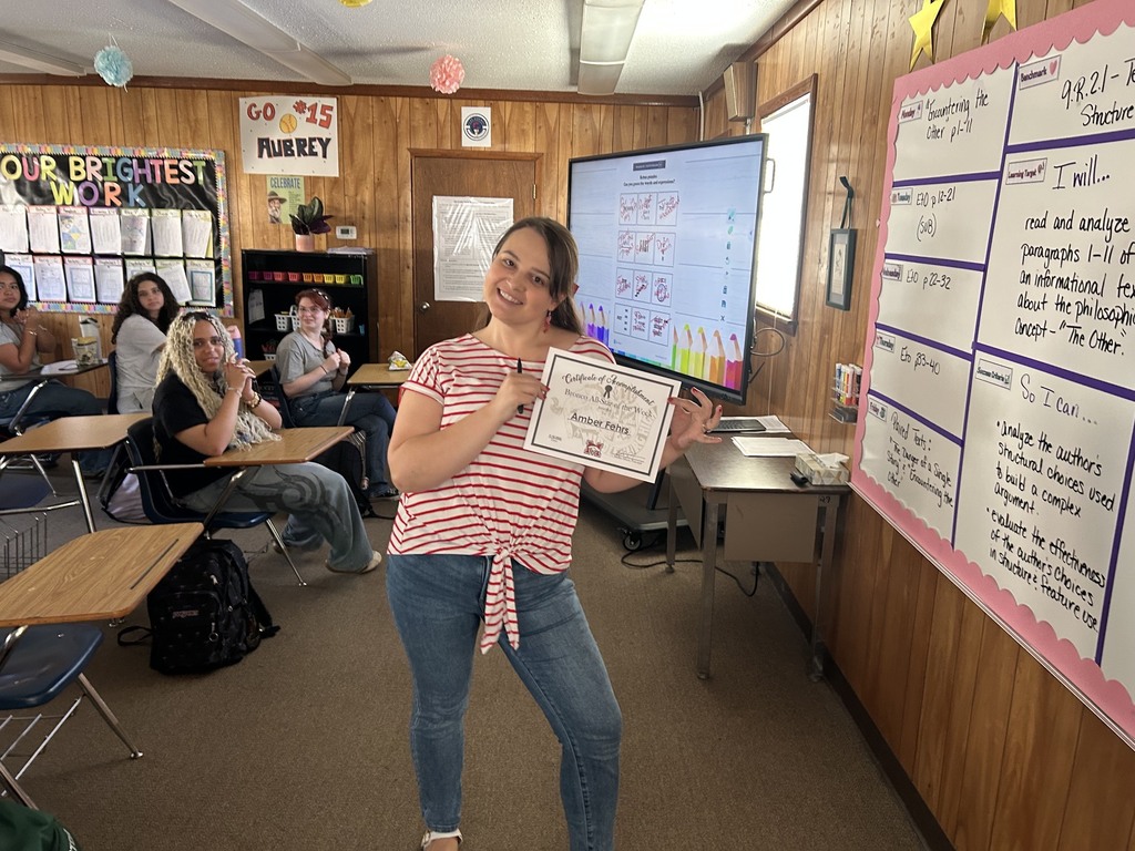 Woman holding certificate standing in front of a classroom, where other students sit at desks.