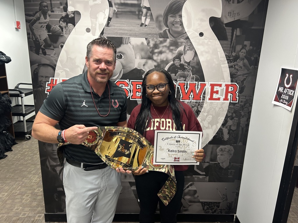 A man and a woman stand together, displaying a championship belt and a certificate in front of a sports-themed wall.