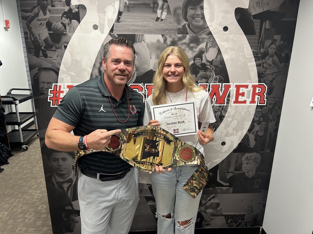 A man and a woman pose with a championship belt, standing in front of a mural featuring sports images.