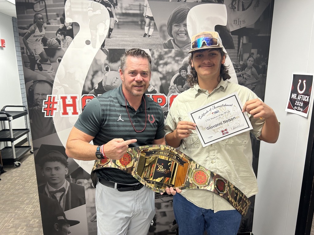 Two people stand next to a large poster. One holds a certificate, and the other has a championship belt.