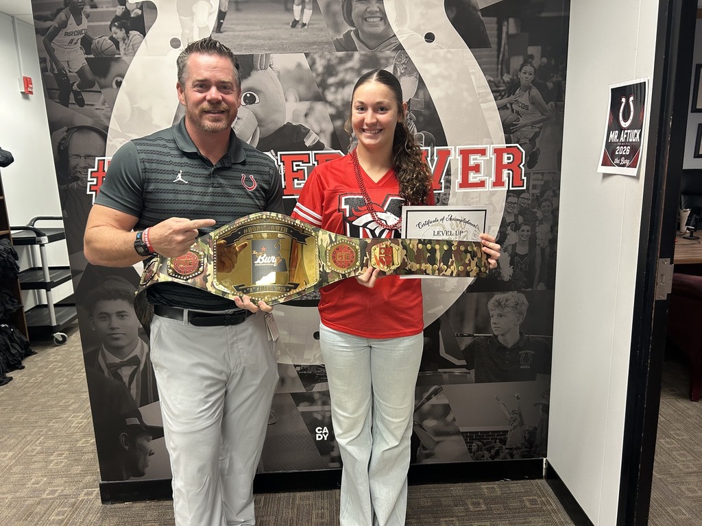 A man and a woman stand, holding a belt. The woman holds a certificate. They are in front of a sports-themed wall.