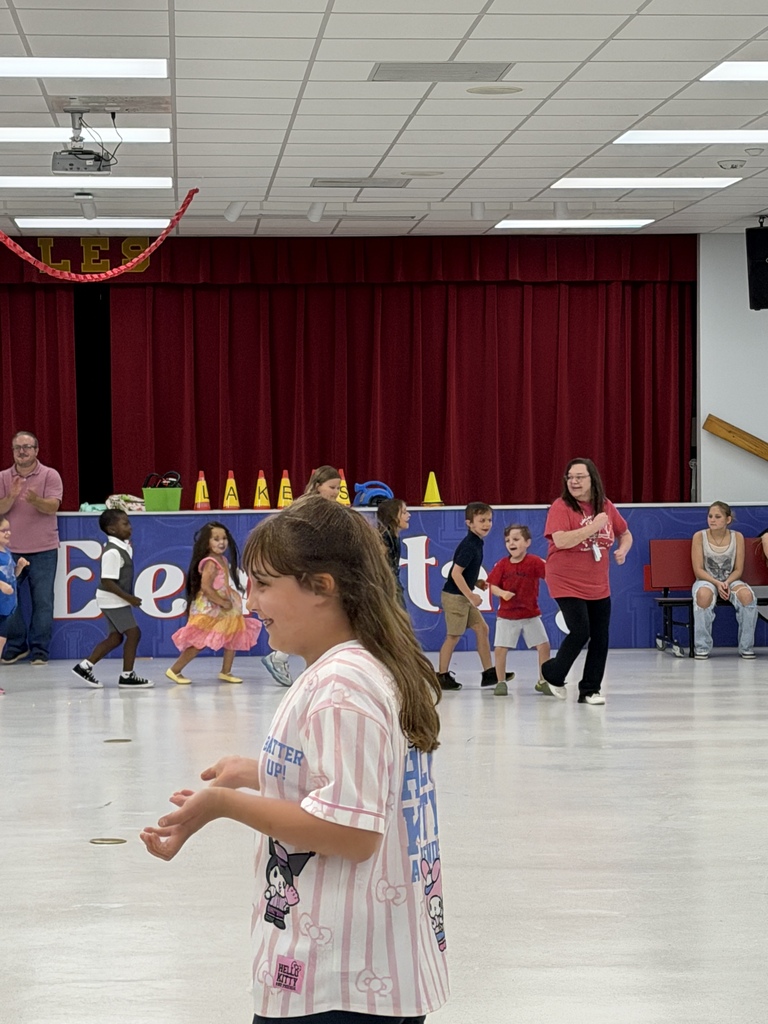 Children in a gymnasium wearing casual clothing. One girl is standing and watching others play a game.