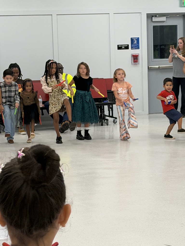 A group of children dance in a room with white walls and a white floor. A woman films from behind.