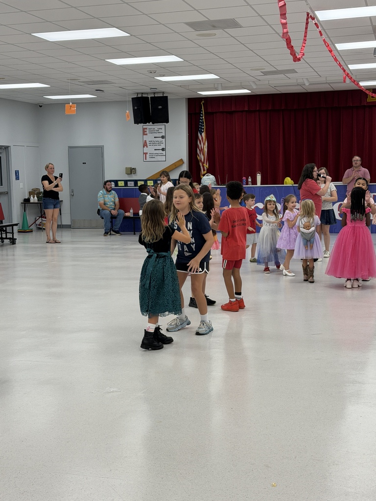 Many children and adults in a room with white floors and a red curtain in the background.