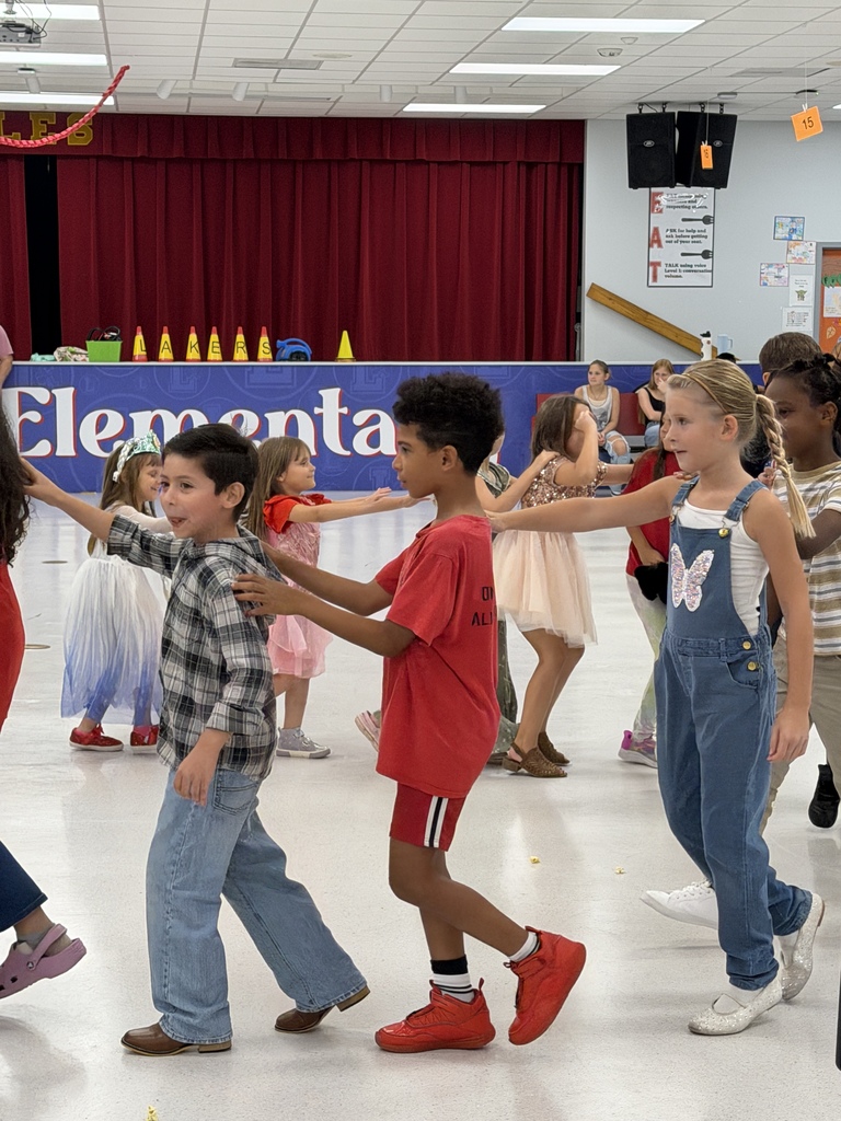 Children dance in a classroom with a banner reading "Elementa". Some wear sneakers, and others wear dresses.