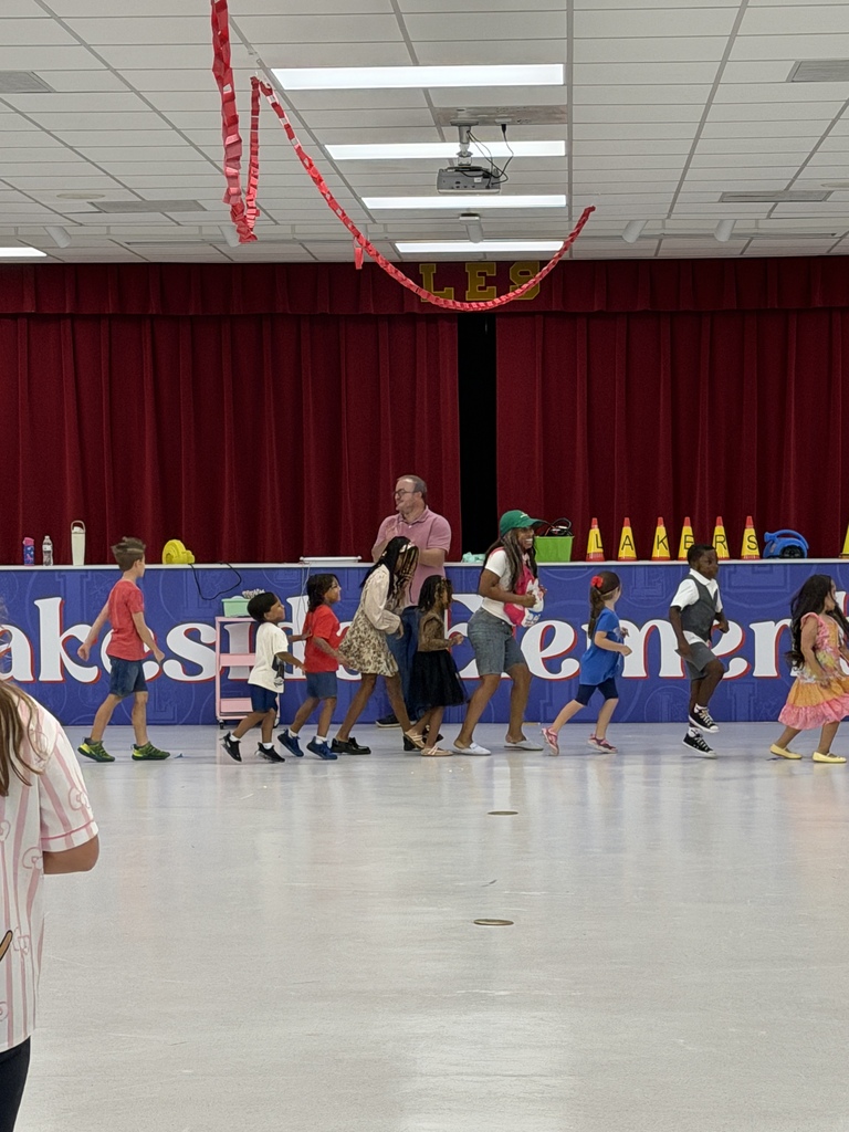 Group of children and adults walking on a floor. Behind them, a long table has bottles. Red curtains hang at the back.