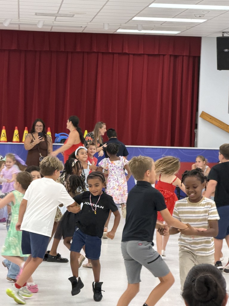 Several children dance in a room with a red curtain and stage, led by adults.