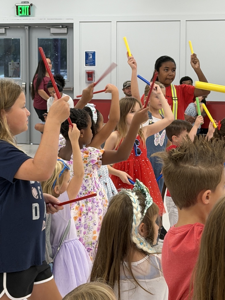 Children in a room with yellow musical instruments, holding them up in the air, with some wearing hats.
