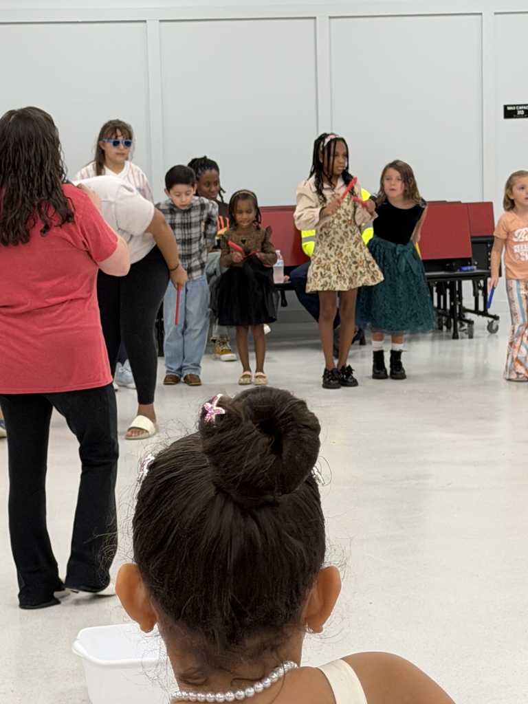 Group of children and adults in a room with a white floor and white walls. Some are standing, others are seated.