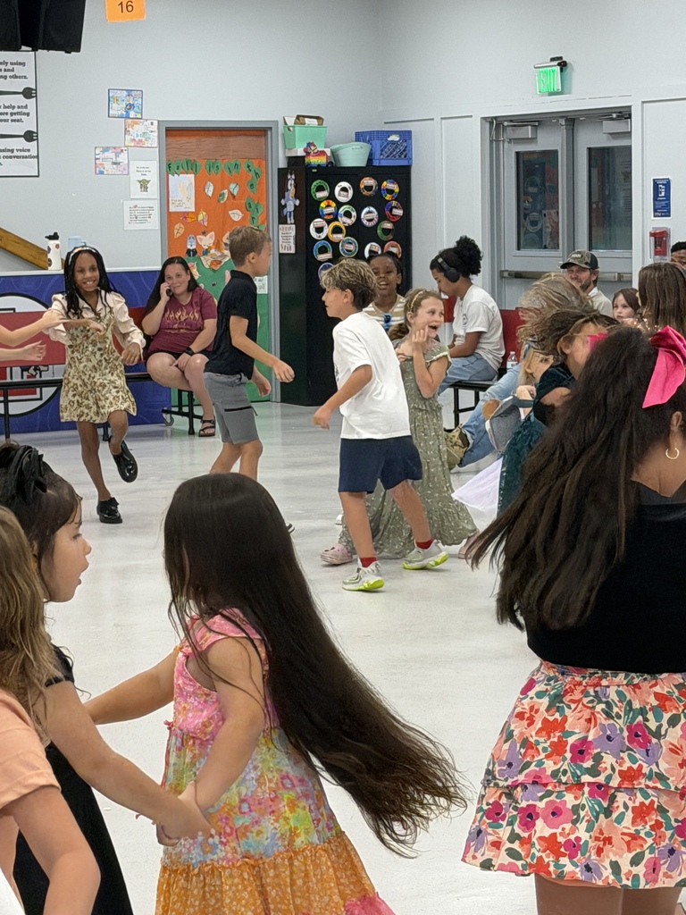 A group of children dancing in a room with white walls. Some adults are seated and observing.