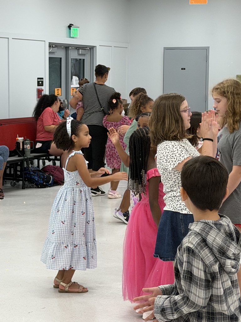 Multiple children in a room, some wearing dresses, dancing and clapping. A person is sitting on a bench nearby.