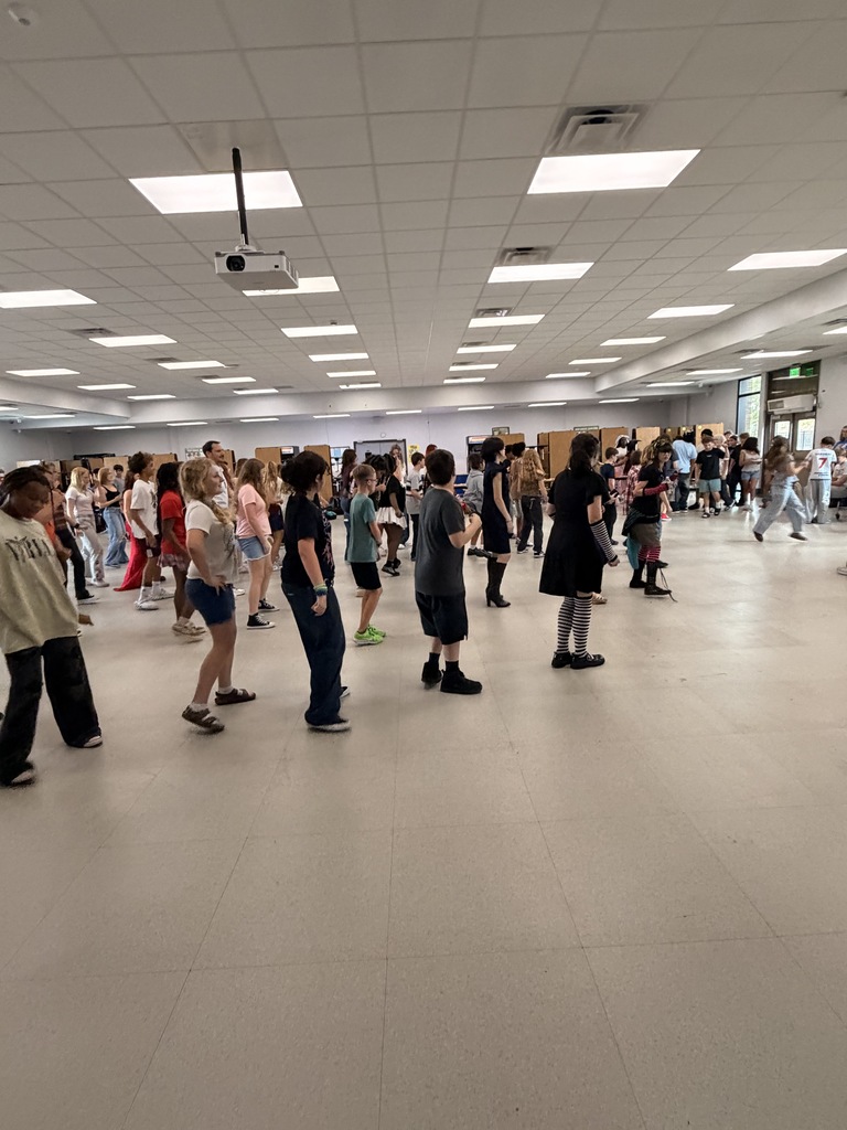 Indoor room with white walls and flooring, ceiling lights, groups of people wearing casual clothing.