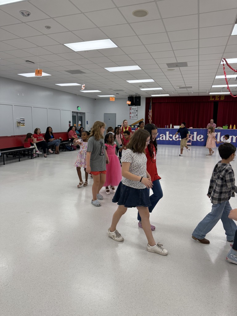 Several children dance inside a room with white floor, red curtains, and people seated along the walls.