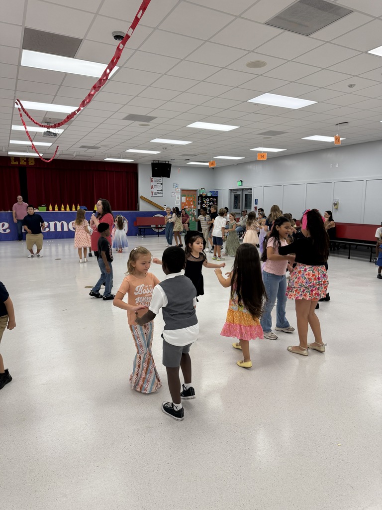 A group of children dance in a large indoor room. A red curtain and ceiling lights are visible.