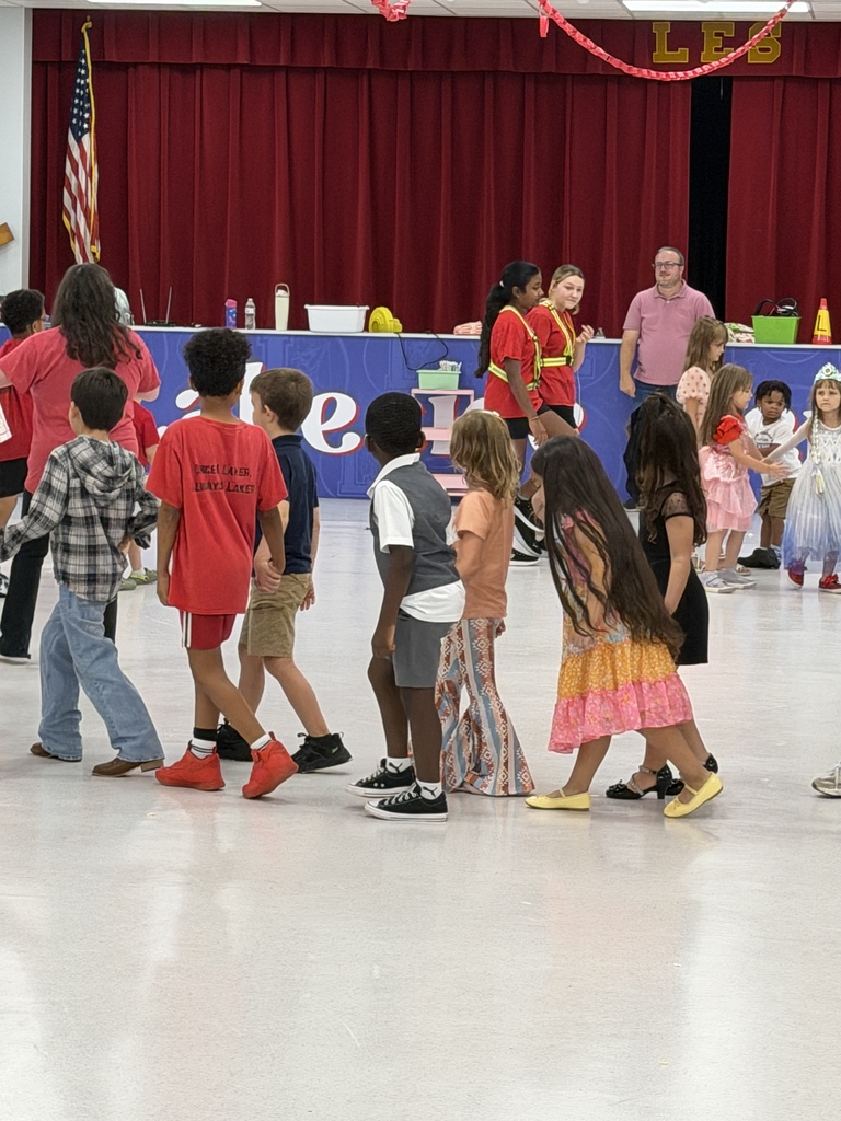 Kids in various outfits stand in a group. Behind them, a table has items on it. A red curtain is in the background.