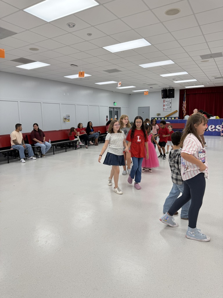 A group of people in a brightly lit indoor area. Some wear sneakers and dance on white floors. Others sit on benches.