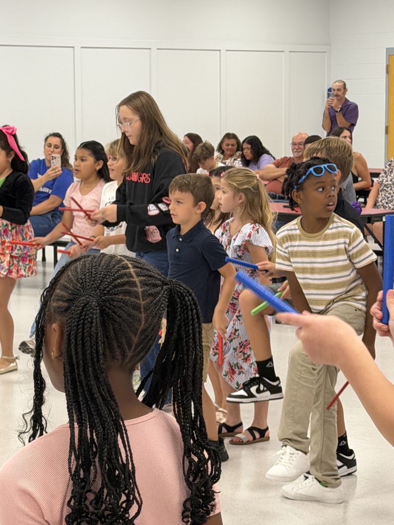 A group of children, some holding musical instruments, are performing in a room with adults observing.