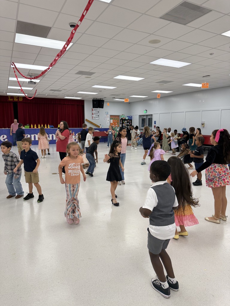 A group of children and adults dance together in a large room with a white floor and stage.