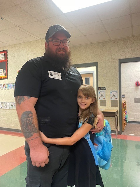 A man with a beard and glasses hugs a young girl in a school hallway.