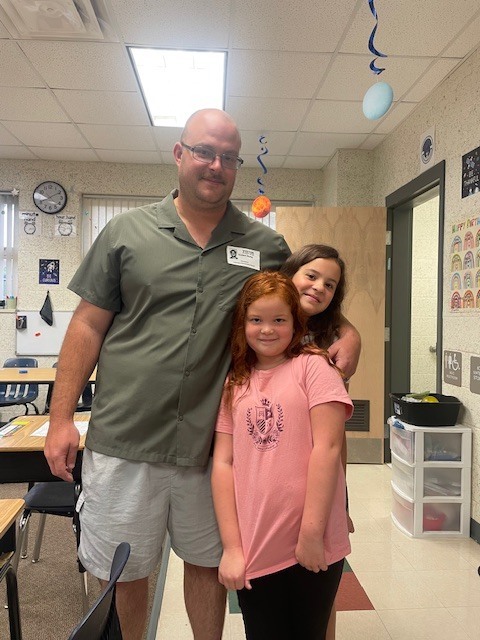Man with glasses stands with two girls in a classroom, posing for a photo.