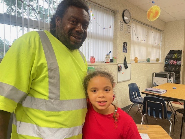 A man in a reflective safety jacket stands next to a young girl in a red shirt in a classroom.