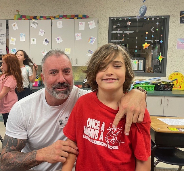 A man and boy smile for a photo. The boy wears a red shirt with white text. Behind them, a classroom is visible with cabinets, desks, and people.