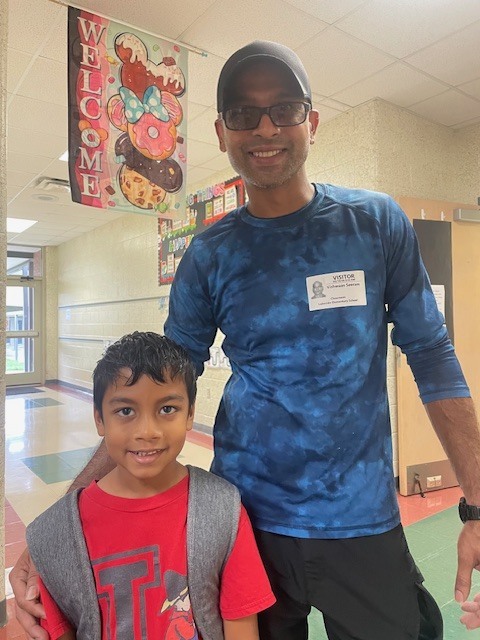A man and a boy stand together in a school hallway. The man wears a blue shirt and black pants. The boy wears a red shirt and a gray vest.