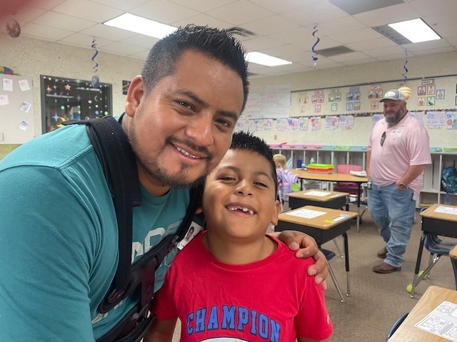 A smiling man in blue stands with a smiling boy in a red shirt, posing for a picture in a classroom.