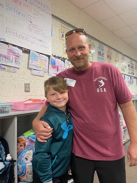 Man in a maroon shirt with "USA" on it standing with a child, smiling. They stand in a classroom with storage boxes, toys, and bulletin boards.