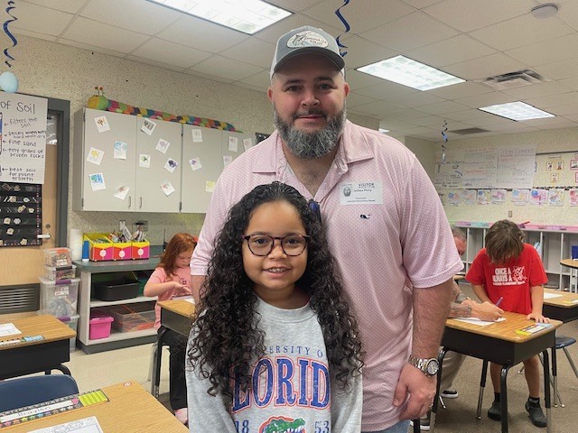 A man with a beard and a hat stands with a smiling girl in glasses in a classroom. Behind them, students sit at desks.