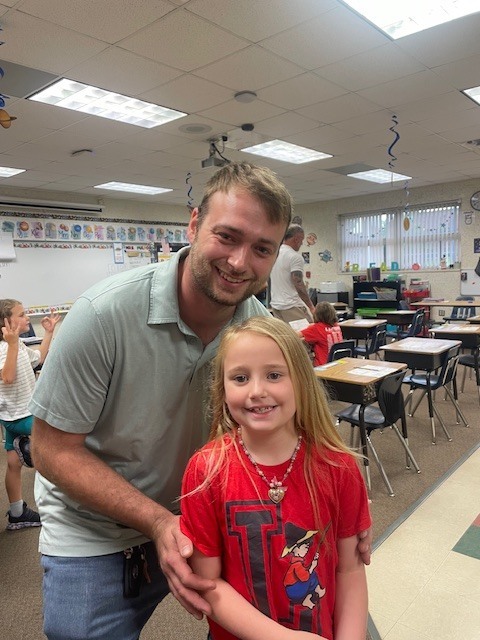 A man and a young girl stand together in a classroom with desks and chairs.