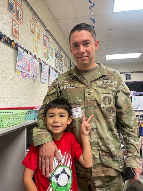 A man in a military uniform stands behind a smiling boy in a red shirt, posing for a photo in a classroom.