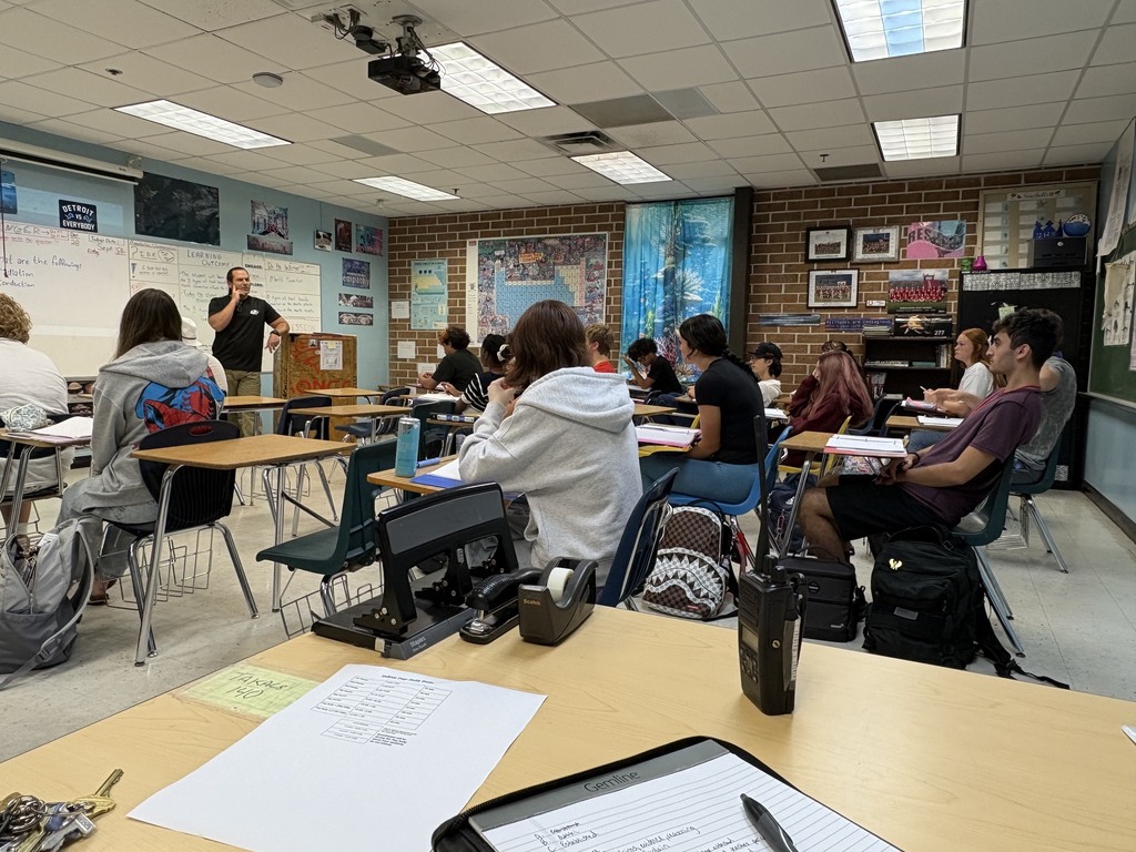 A classroom with desks and chairs, students sitting and a teacher standing in front of a whiteboard.