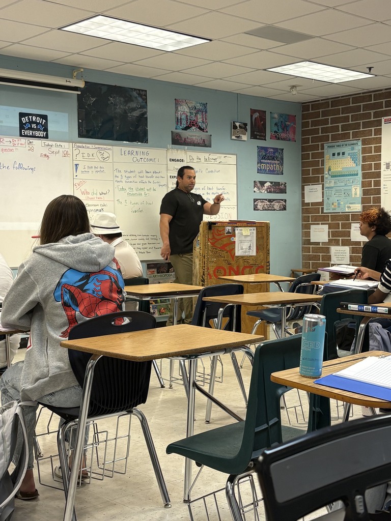 A classroom with desks and chairs. A person stands at a podium, speaking to a seated person.