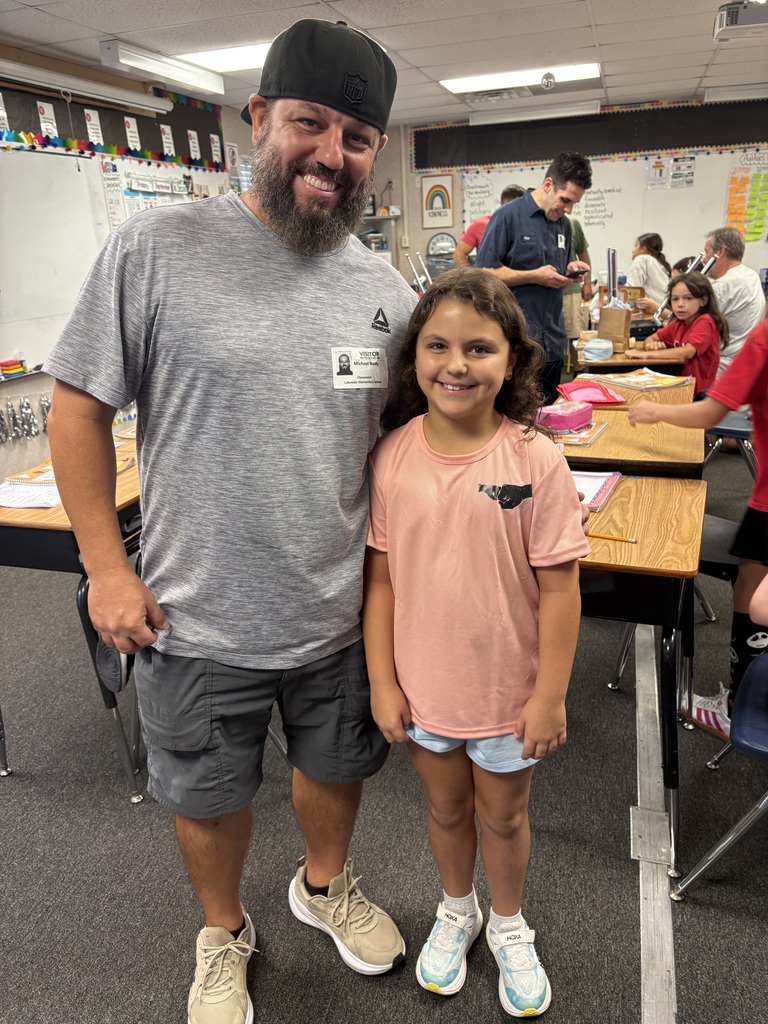 A man and a young girl stand in a classroom, smiling for the camera. Behind them, people are seated at desks.