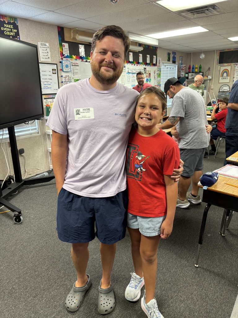 A man and a young girl stand in a classroom with desks and adults working.