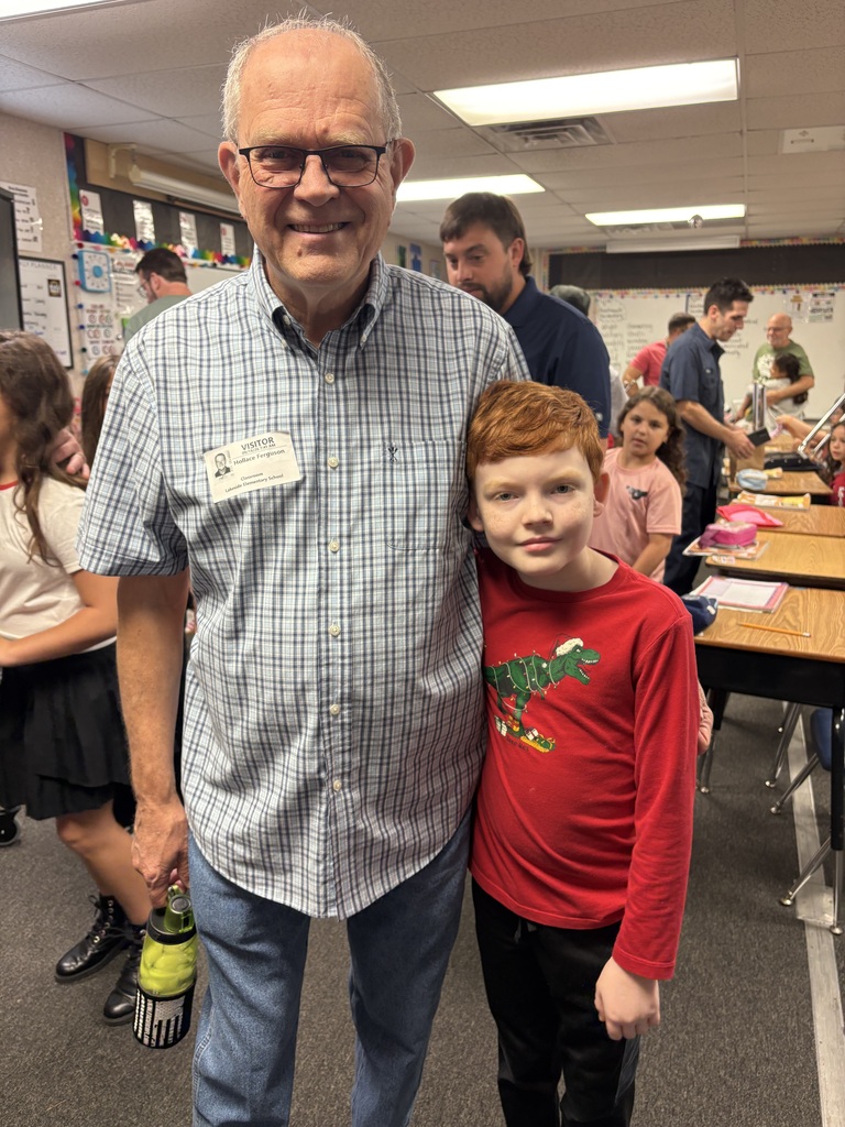 A man in glasses and a checkered shirt stands next to a boy in a red shirt in a classroom.