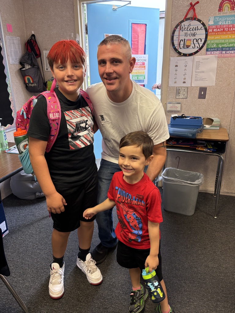 Three children stand in a classroom; one wearing a backpack, another in a red shirt, and a man in a white t-shirt behind them.