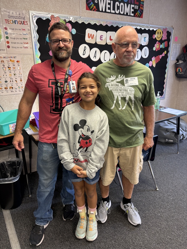 Three people in a classroom, a young girl in a sweatshirt, and two men in casual wear. Behind them is a bulletin board with the words "Welcome Back".