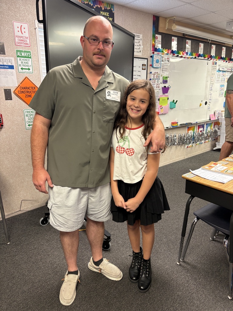 Adult male wearing glasses stands next to a young girl, both smiling. They are in a classroom with a white board, desks, and a dark monitor in the background.