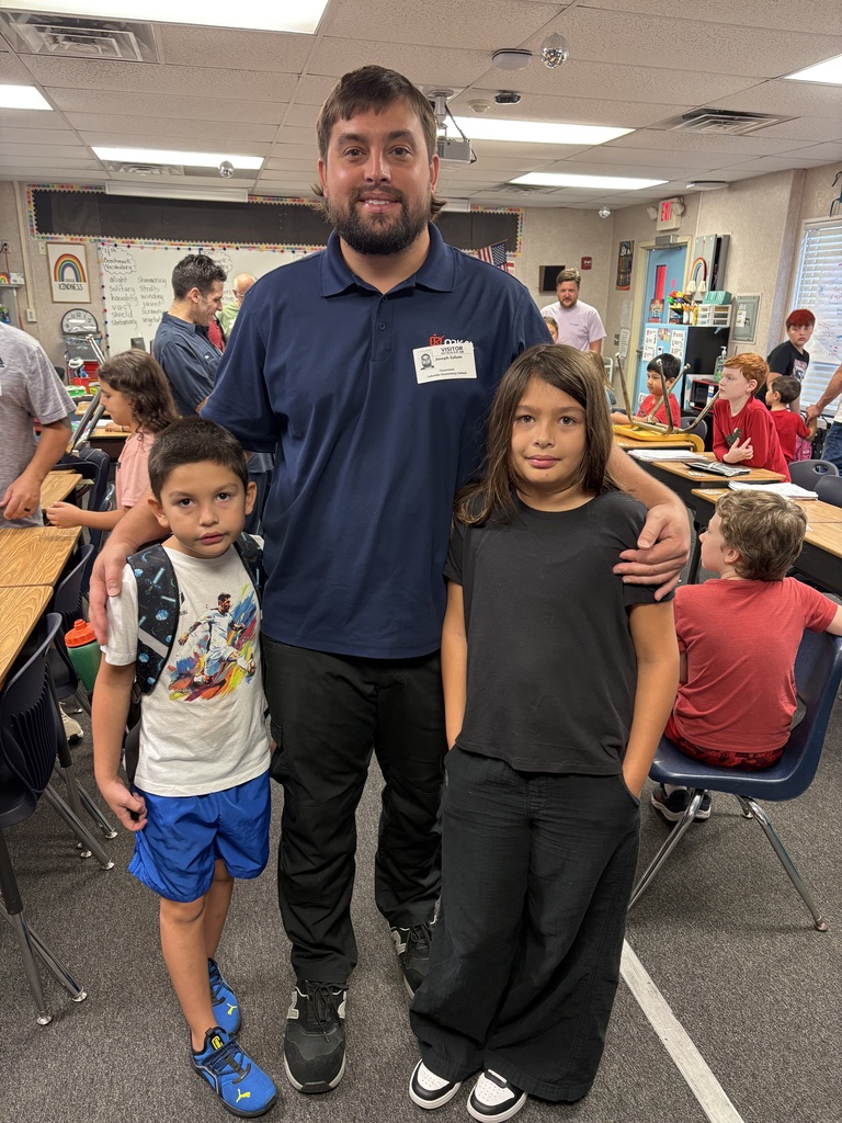 A man in a blue shirt stands between two children in a classroom with desks and other students.