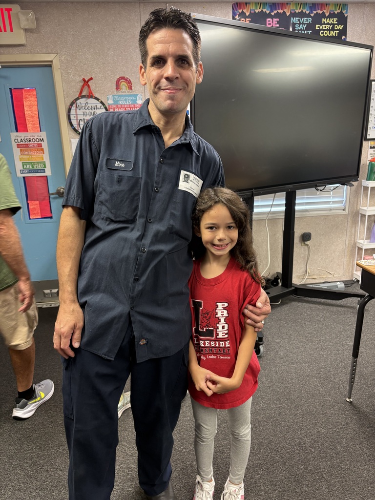 A man in a navy blue shirt and a young girl in a red shirt stand close together. Behind them, a large monitor and shelves are visible.