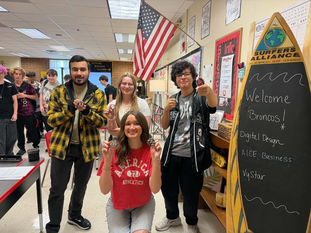 Five people stand in a classroom, smiling. Two large posters with text are behind them. An American flag hangs on the wall.